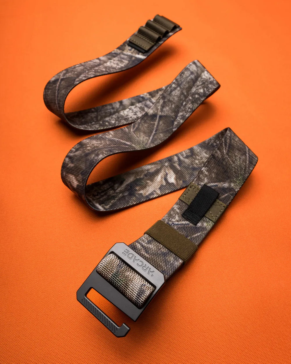 A camouflage-patterned nylon belt with a metal buckle and black rectangular accent is displayed on an orange surface. The belt is loosely coiled, showing its design and texture.