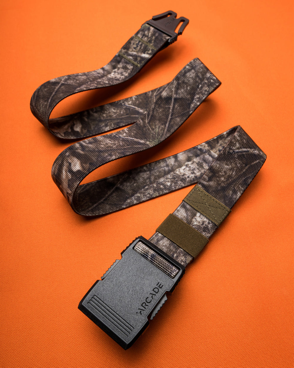 A camouflage-patterned stretch webbing belt with a low profile plastic buckle labeled ARCADE is laid out on an orange textured surface.
