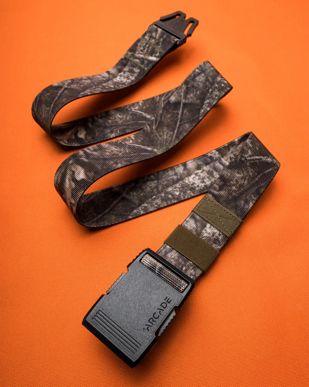 A camouflage-patterned fabric belt with a black plastic buckle labeled “ARCADE” is arranged on an orange background.