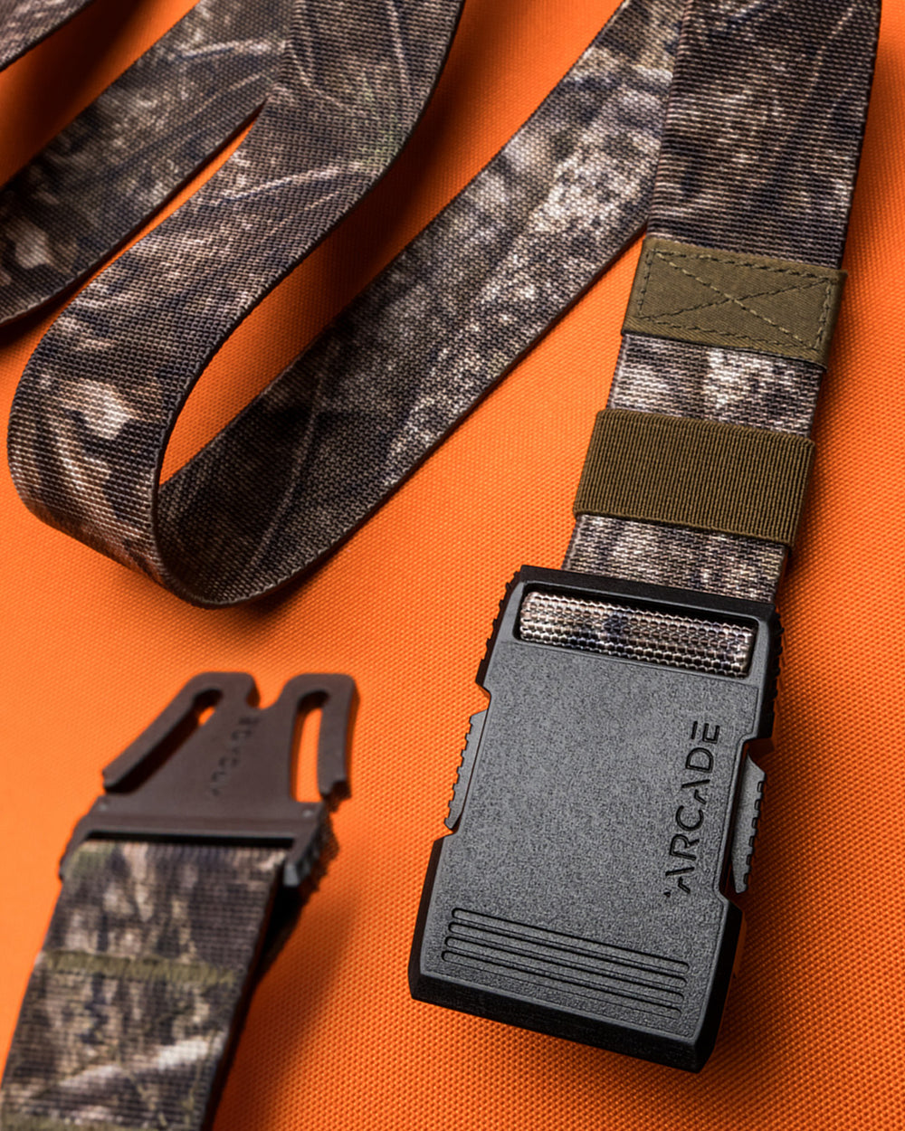 A close-up of a camouflage-patterned Arcade Belt with stretch webbing and a black buckle, laid out on an orange textured surface. The buckle is partially detached, revealing the belt’s ARCADE branding.