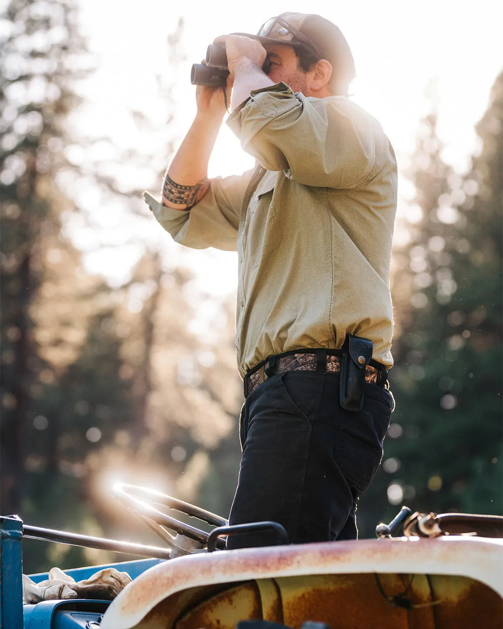 A person in a green shirt and dark pants stands on a blue vehicle outdoors, looking through binoculars. Sunlight filters through tall trees in the background, creating a warm, natural setting.