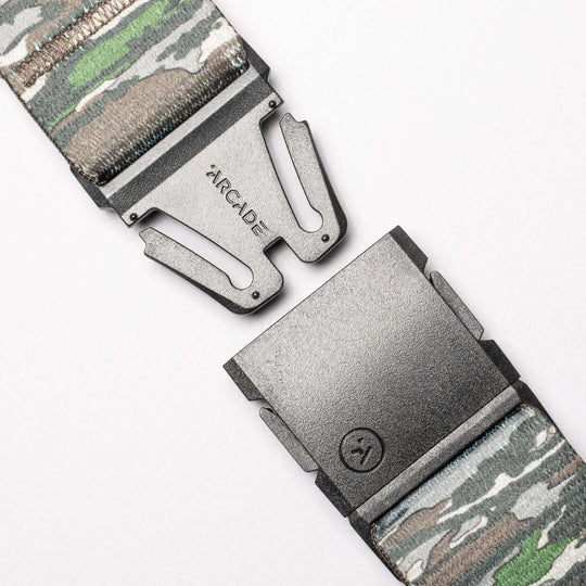 Close-up of the Realtree Camo Original by Arcade Belts, a stretch webbing belt with a camouflage pattern, partially open metal buckle engraved “ARCADE,” and a small lower logo.
