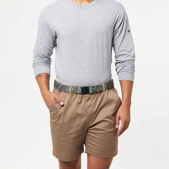 Someone stands against a plain white background wearing a light grey long-sleeve shirt, brown shorts, and the Arcade Belts Realtree Kit low profile elastic stretch belt with hands in pockets.