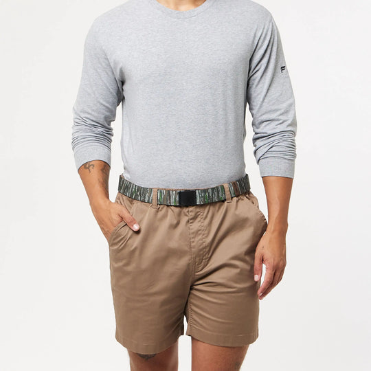 Someone stands against a plain white background wearing a light grey long-sleeve shirt, brown shorts, and the Arcade Belts Realtree Kit low profile elastic stretch belt with hands in pockets.