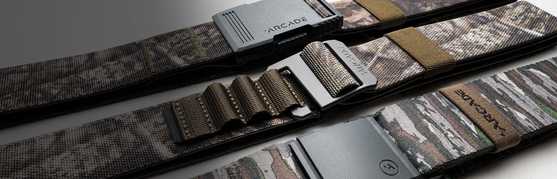 Three camouflage-patterned Arcade brand Low Profile Belts with different plastic buckles and elastic stretch fit are laid out side by side on a white surface, partially in shadow.