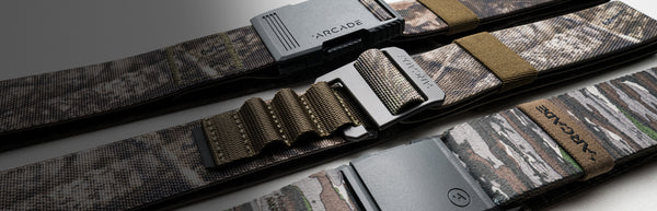 Three camouflage-patterned Arcade brand Low Profile Belts with different plastic buckles and elastic stretch fit are laid out side by side on a white surface, partially in shadow.