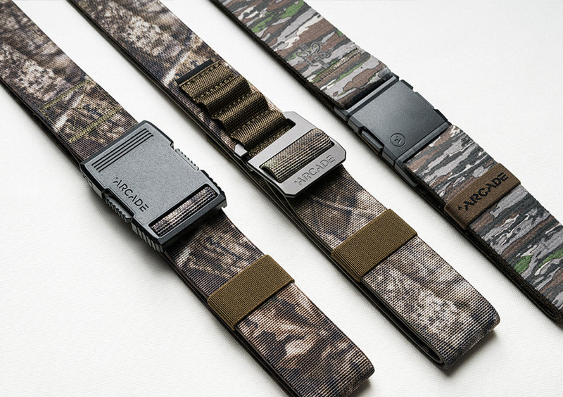 Three Arcade Low Profile Belts with unique camo designs and different buckles—black, silver, and dark gray—are laid out parallel on a white surface. Each features durable stretch webbing for comfort and flexibility.