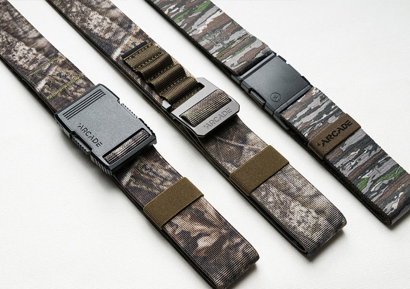 Three camouflage-patterned Arcade brand travel belts, made with stretch webbing and featuring different buckle styles, are laid out side by side on a white surface.