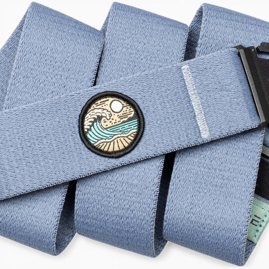 The Arcade Belts Save the Waves belt features a light blue surf-ready design, a round wave-sun-sand patch, black buckle, stitched white line detail, and supports conservation efforts inspired by Save the Waves.