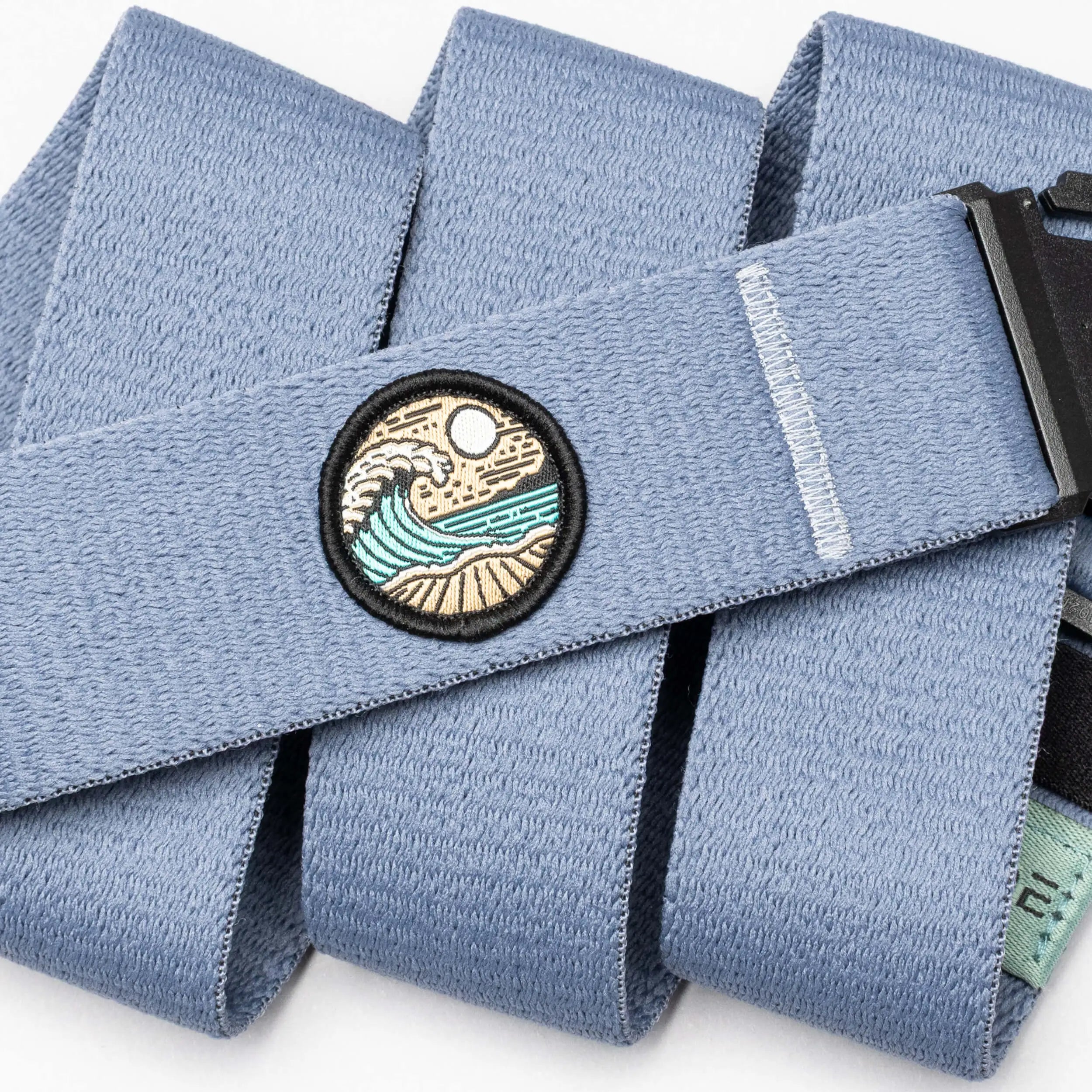 The Arcade Belts Save the Waves belt features a light blue surf-ready design, a round wave-sun-sand patch, black buckle, stitched white line detail, and supports conservation efforts inspired by Save the Waves.