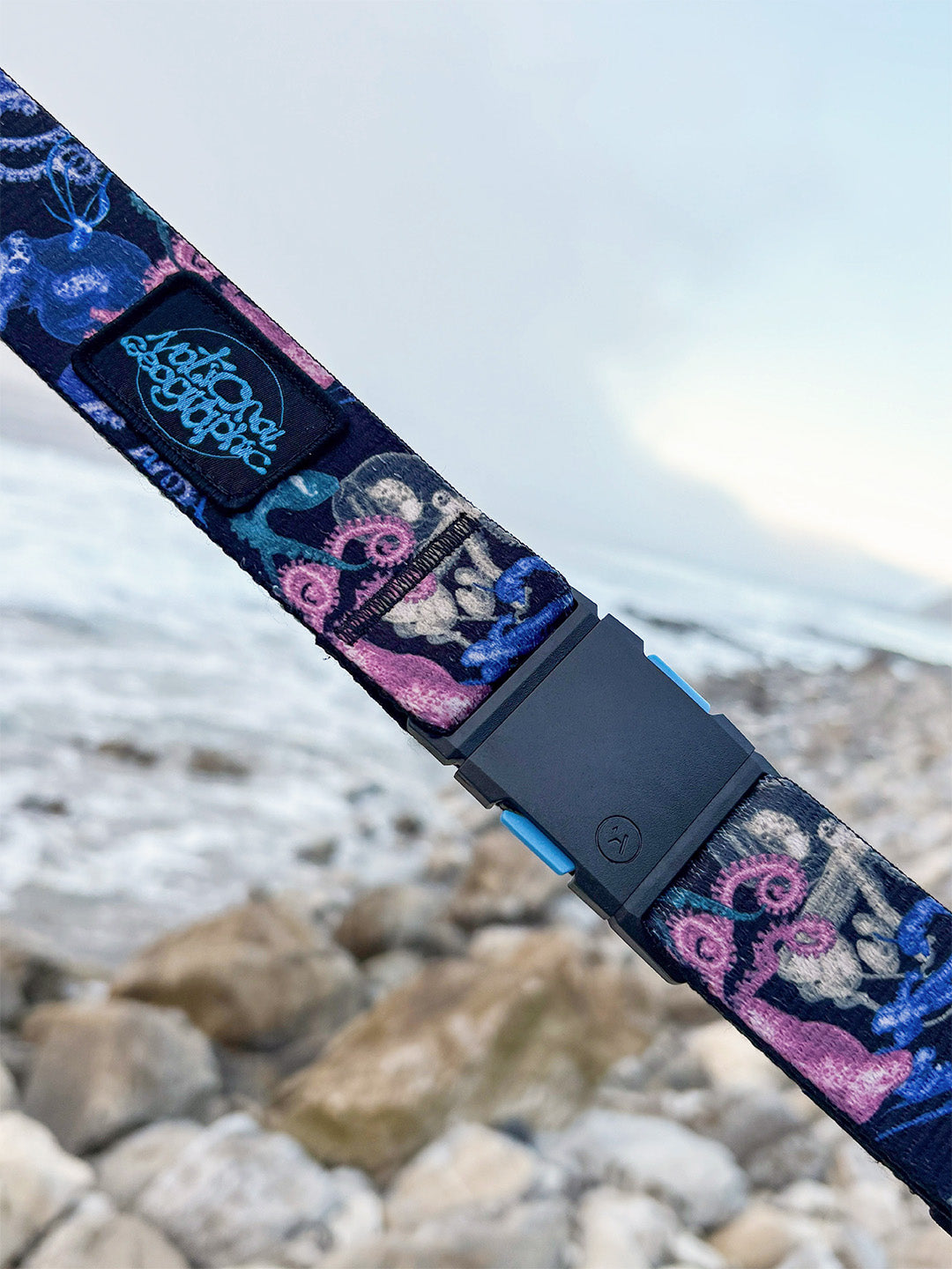 A colorful, patterned belt with a black buckle is held in front of a rocky shoreline and an overcast, cloudy sky by the ocean. The belt features designs in blue, pink, and cream tones.