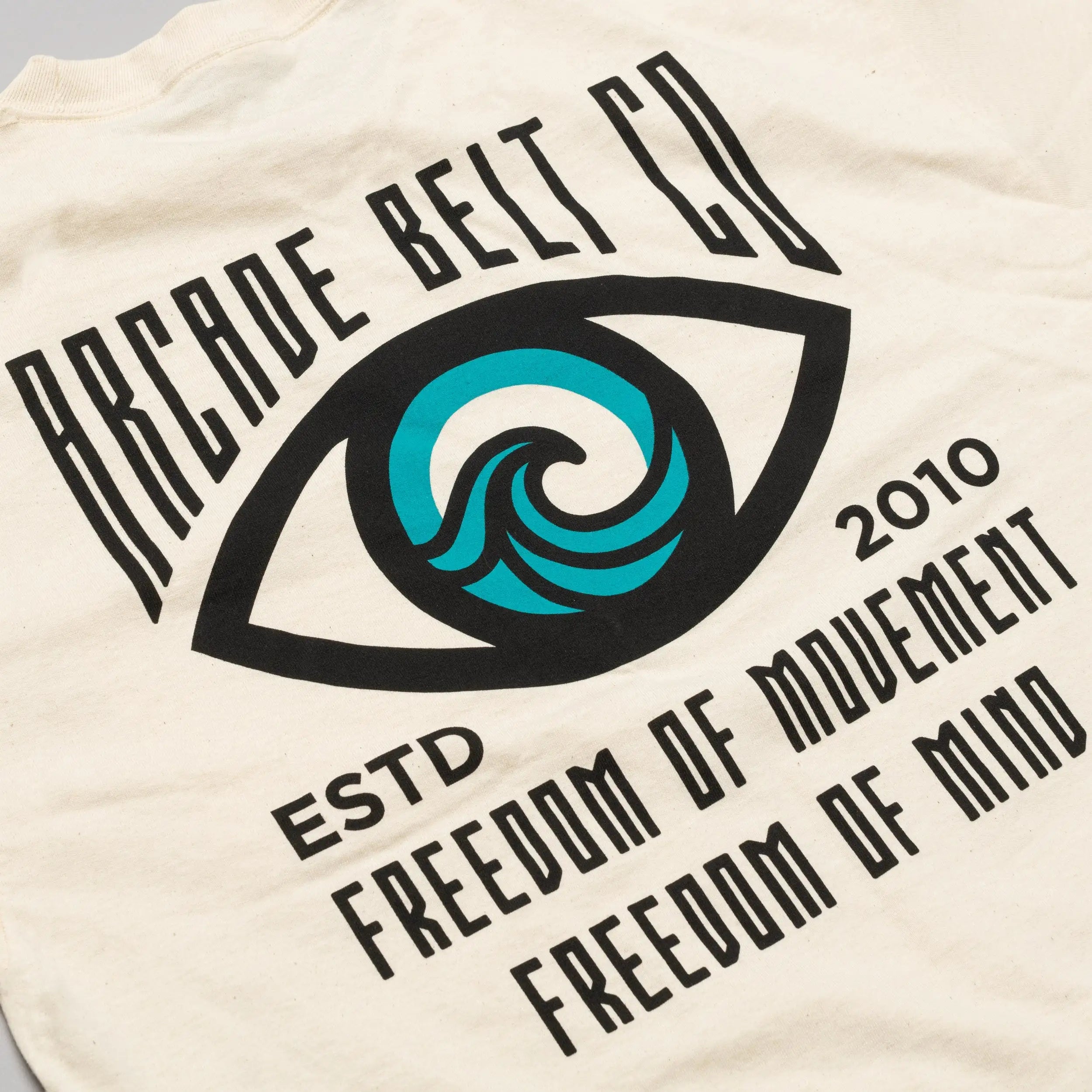 The Arcade Freedom Tee by Arcade Belts is a cream-colored shirt with a large eye graphic featuring a blue wave and text: Arcade Belt Co, ESTD 2010, Freedom of Movement, and Freedom of Mind—ideal for comfort enthusiasts.