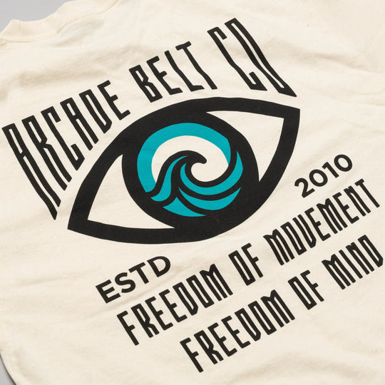 The Arcade Freedom Tee by Arcade Belts is a cream-colored shirt with a large eye graphic featuring a blue wave and text: Arcade Belt Co, ESTD 2010, Freedom of Movement, and Freedom of Mind—ideal for comfort enthusiasts.