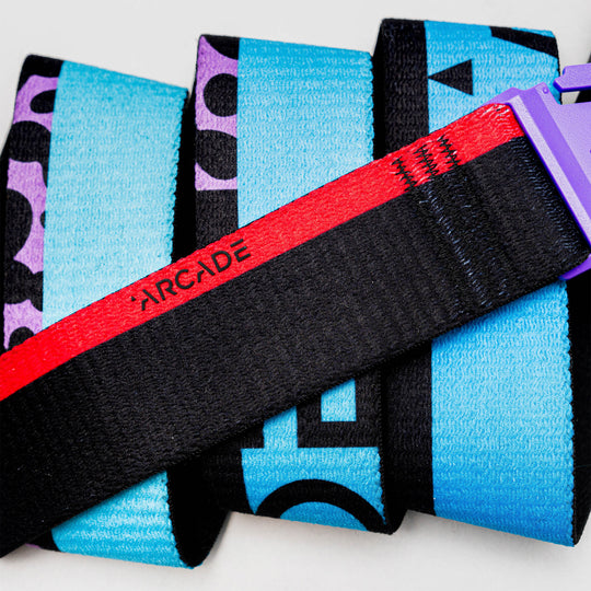 A close-up of the Sierra black Travel Belt by Arcade Belts, featuring a red stripe, purple plastic buckle, and ARCADE text, laid on blue and purple patterned fabric with bold black letters.