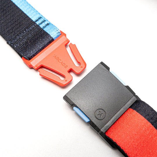 A close-up of Arcade Belts Sierra Elastic Stretch Belt, showing its red ARCADE-branded plastic buckle at one end and a black rectangular buckle with blue accent on the other—perfect as a travel belt.