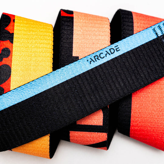 A close-up of the Arcade Belts Sierra Elastic Stretch Belt, showcasing its colorful textured design with a blue and black center and the word ARCADE, set against a background of red, orange, and yellow abstract patterns.