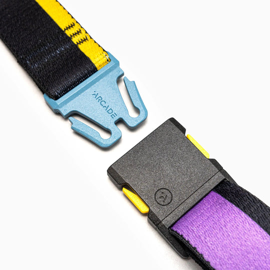 Close-up of the Arcade Belts Sierra Elastic Stretch Belt with a blue metal buckle marked ARCADE and black plastic clasp, featuring black, yellow, and purple woven straps on a white background.