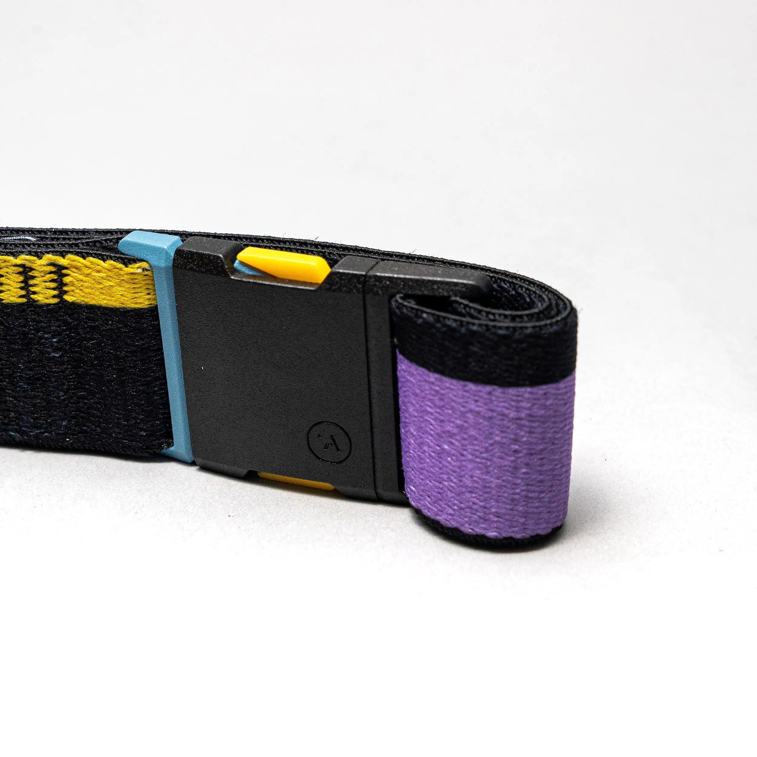 A close-up of the Arcade Belts Sierra: a rolled-up black elastic stretch belt with a purple end, yellow and blue accents, and a black rectangular buckle with a small triangle logo, set against a plain light background.