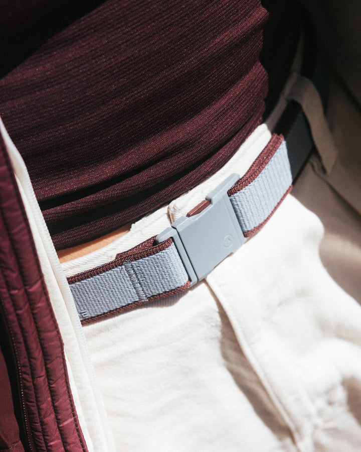 Close-up of a person wearing a maroon shirt, white pants, and a gray stretch webbing low profile belt with a plastic buckle. Sunlight casts shadows, highlighting the durable travel belt and the textured fabric of the shirt.