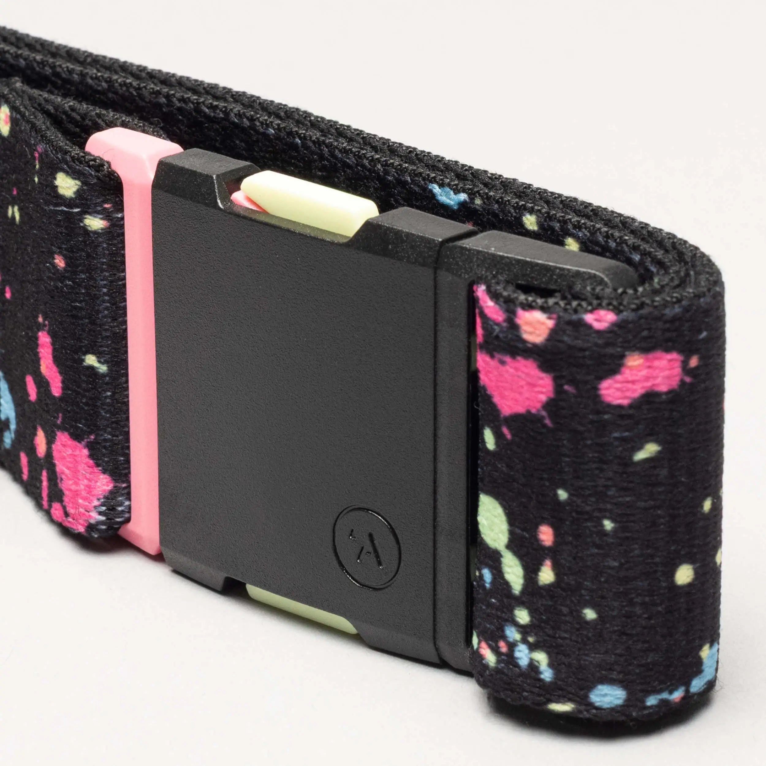 A close-up of the Splatter belt by Arcade Belts shows a black plastic buckle with a circular logo. The fabric pops with a drip-inspired print—pink, green, and blue spots on black—for extra chill style.