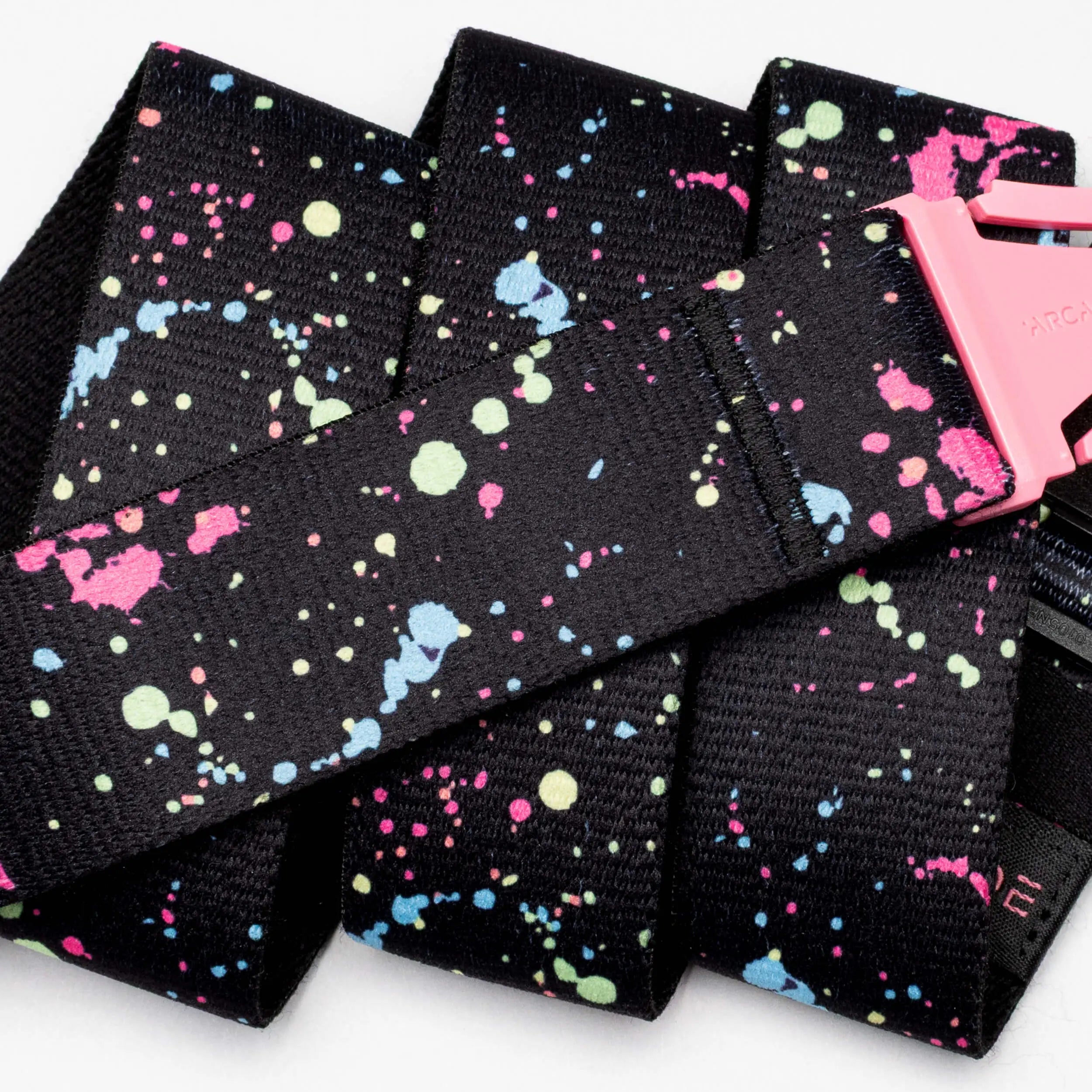 The Arcade Belts Splatter features a black strap with a pink buckle and colorful paint drip splatters in blue, pink, yellow, and green, shown in overlapping loops against a white background.