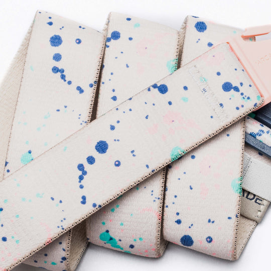 A close-up of the Arcade Belts Splatter fabric resistance band, set against a cream background with blue, teal, and pink paint splatter patterns. The band features stitched edges and a rectangular rubber label on one end.