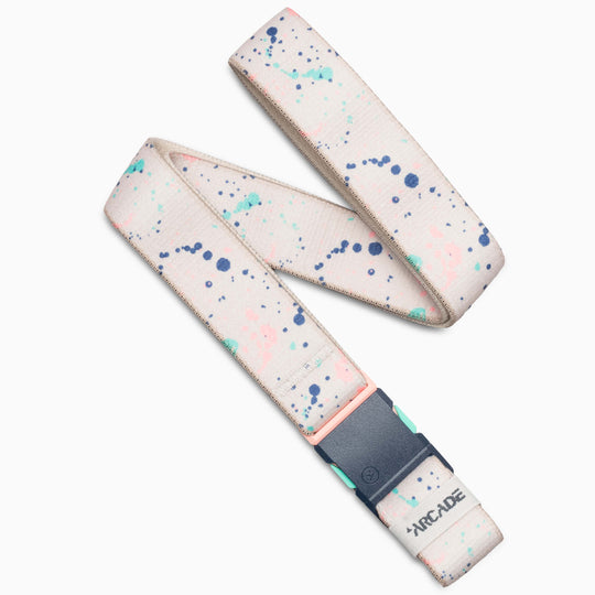 The Arcade Belts Splatter is a beige fabric belt with blue, green, and pink paint splatters, a dark gray buckle, and a small ARCADE logo near the end. It is displayed on a white background.