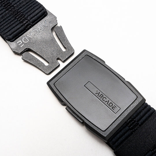 A close-up of a black Arcade Belts Stamped Travel Belt with a plastic buckle, displaying the brand name on the buckle and an elastic stretch webbing strap, set against a white background.