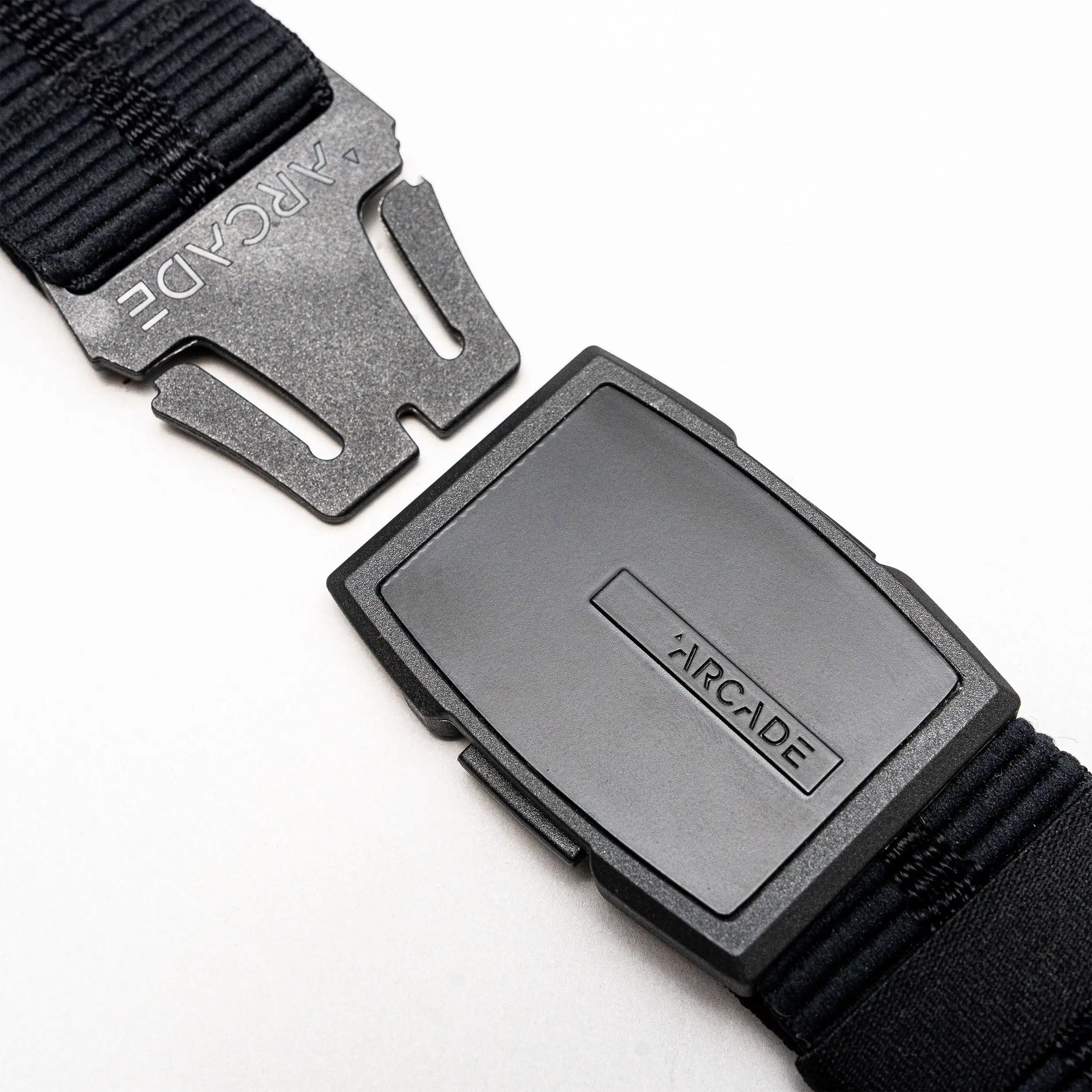 A close-up of a black Arcade Belts Stamped Travel Belt with a plastic buckle, displaying the brand name on the buckle and an elastic stretch webbing strap, set against a white background.