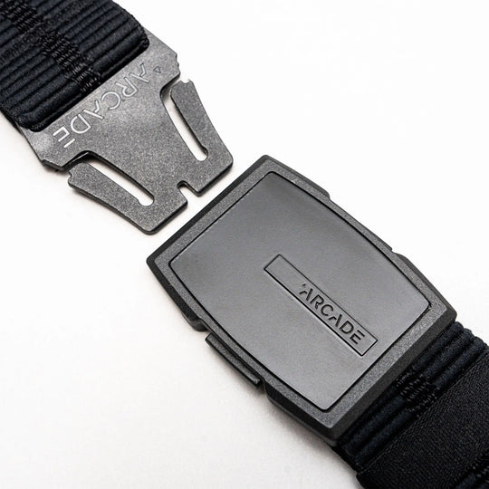 A close-up of a black Arcade Belts Stamped Travel Belt with a plastic buckle, displaying the brand name on the buckle and an elastic stretch webbing strap, set against a white background.