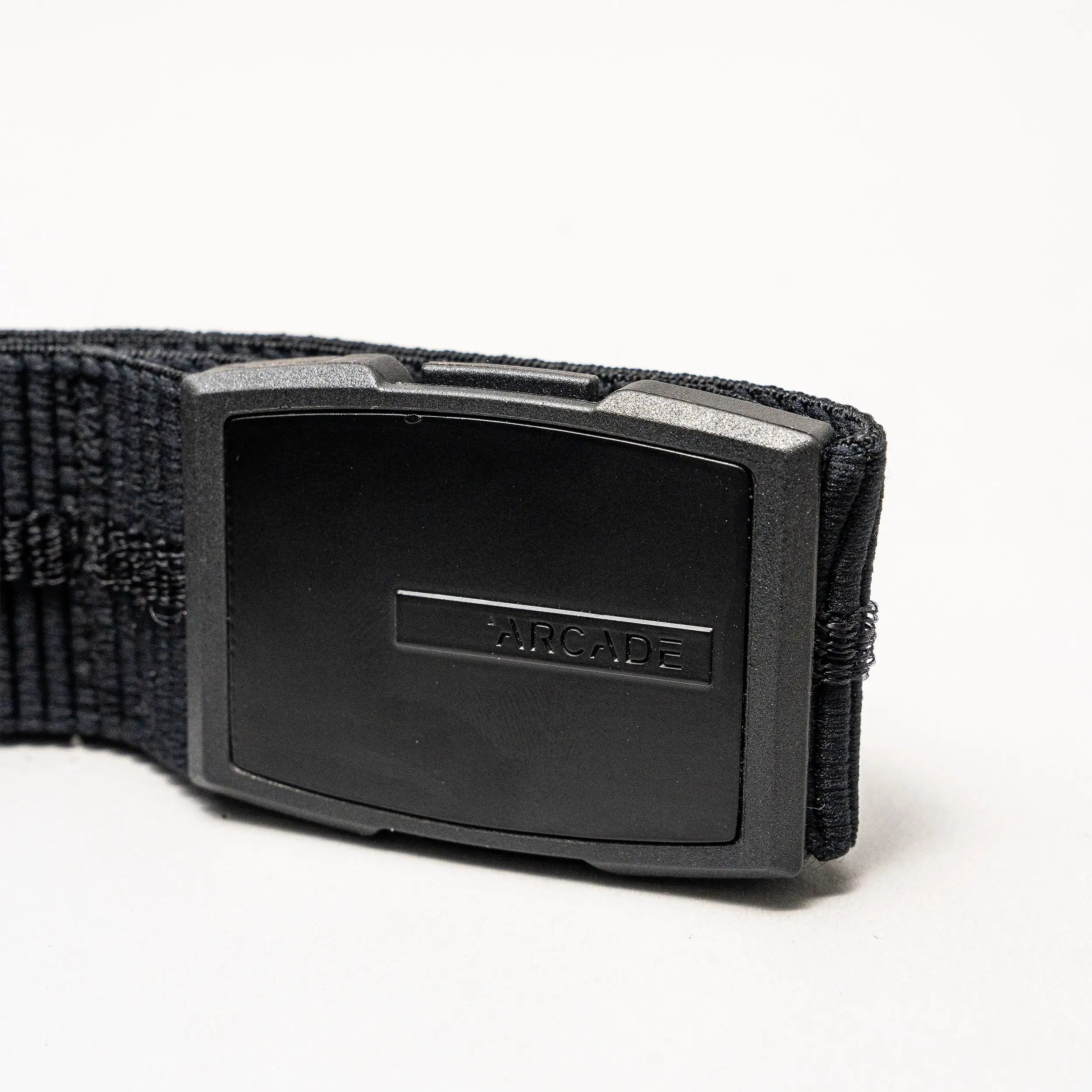 A close-up of the Arcade Belts Stamped, a low-profile belt with a rectangular black plastic buckle embossed with ARCADE. Its stretch webbing ensures comfort. The product is shown against a plain white background.