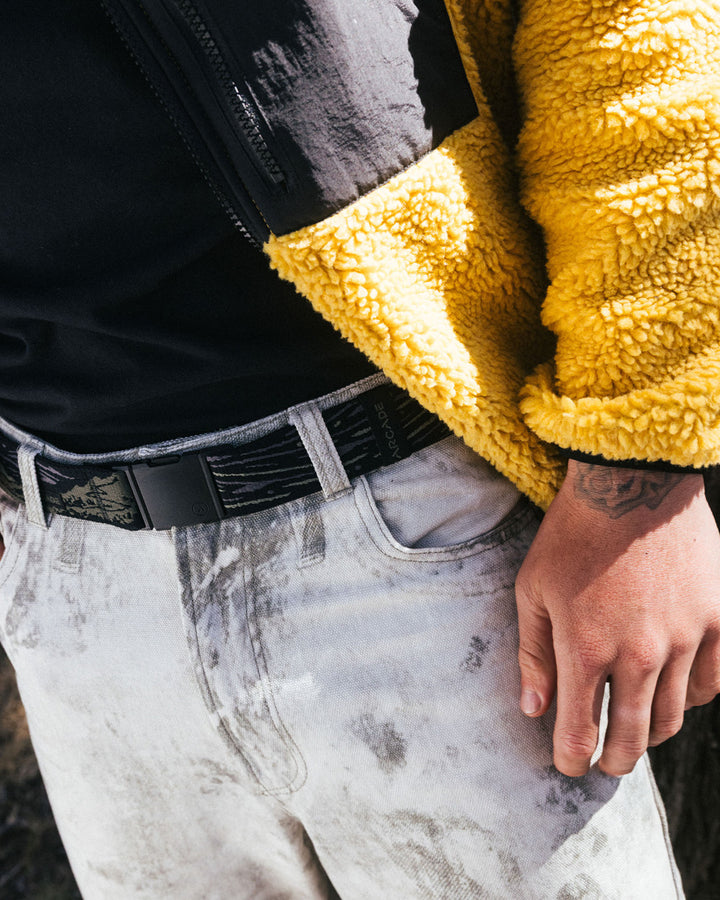 A person wearing a yellow sherpa jacket, black shirt, and light-washed jeans with a travel belt featuring a plastic buckle, showing only their torso and hand.