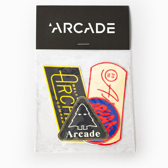 The Arcade Sticker Pack by Arcade Belts includes four colorful stickers—a yellow rectangle, a round sticker, an oval sticker, and a black triangle with a tree and “Arcade Belts”—all in a plastic bag labeled Arcade.