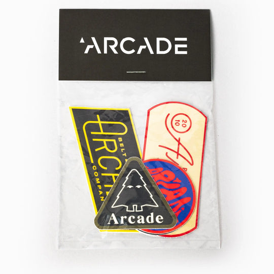 The Arcade Sticker Pack by Arcade Belts includes four colorful stickers—a yellow rectangle, a round sticker, an oval sticker, and a black triangle with a tree and “Arcade Belts”—all in a plastic bag labeled Arcade.
