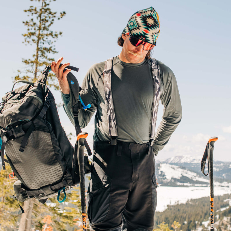 A person wearing sunglasses, a colorful knit hat, and outdoor gear—including a low profile belt—holds a backpack and hiking poles while standing in a snowy mountain landscape under a clear sky.