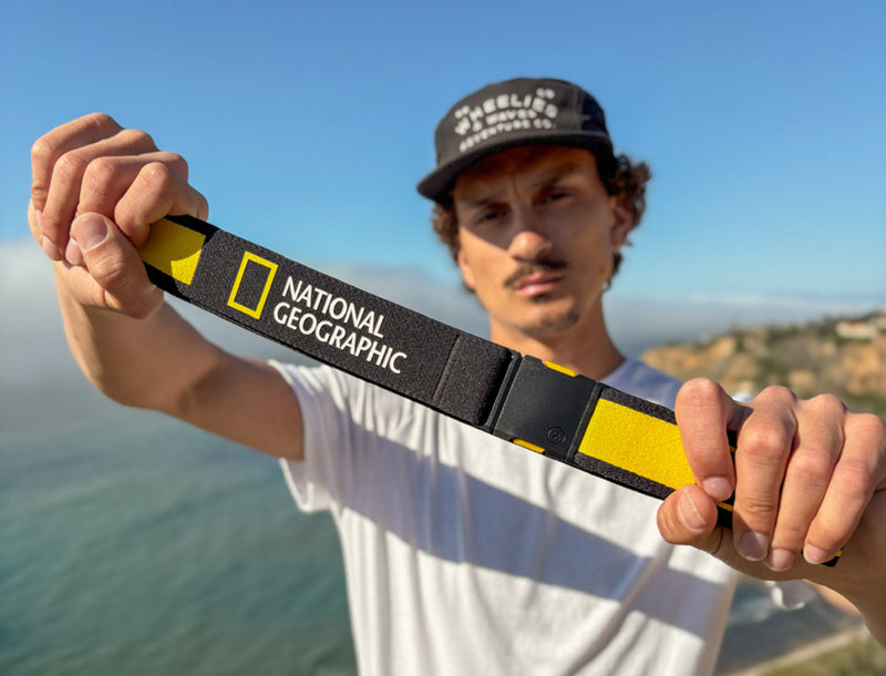 A man in a white t-shirt and black cap holds out a black and yellow strap with the National Geographic logo, standing outdoors near a body of water and cliffs under a clear sky.