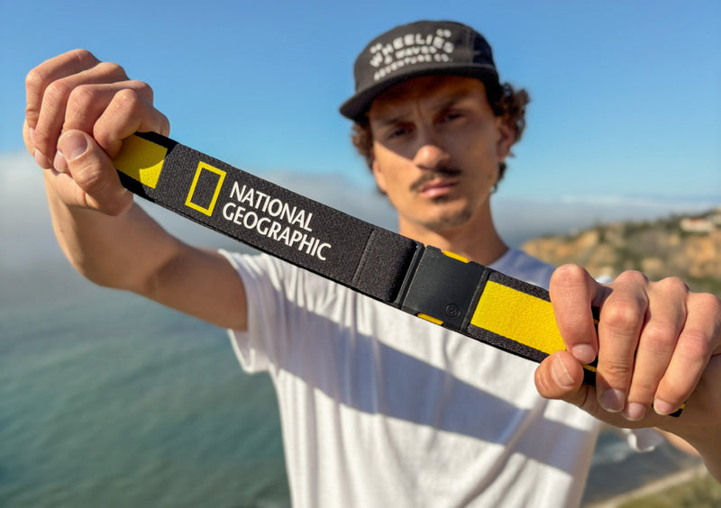 A man in a white shirt and black cap holds a camera strap with the National Geographic logo, standing outdoors near the ocean with cliffs in the background on a sunny day.