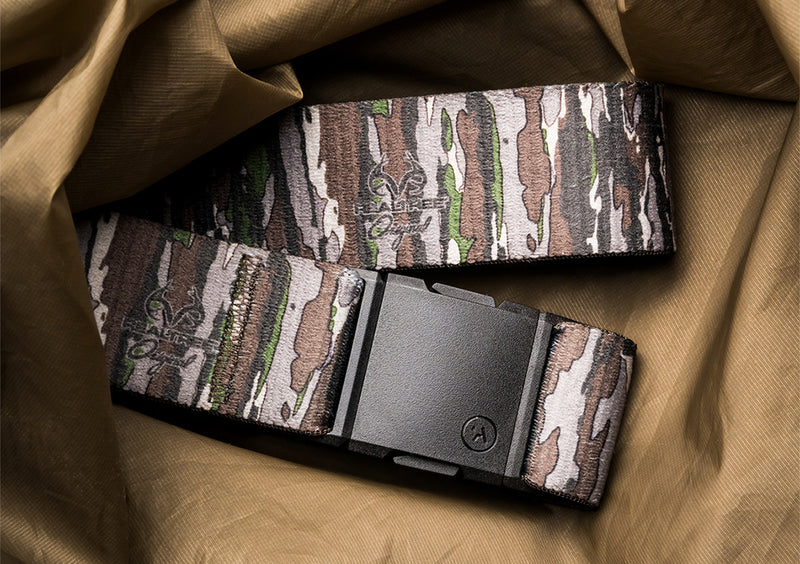 A camouflage-patterned stretch webbing belt with a black buckle lies on a crumpled khaki fabric background, perfect for those seeking a comfortable and low profile belt for travel or everyday wear.