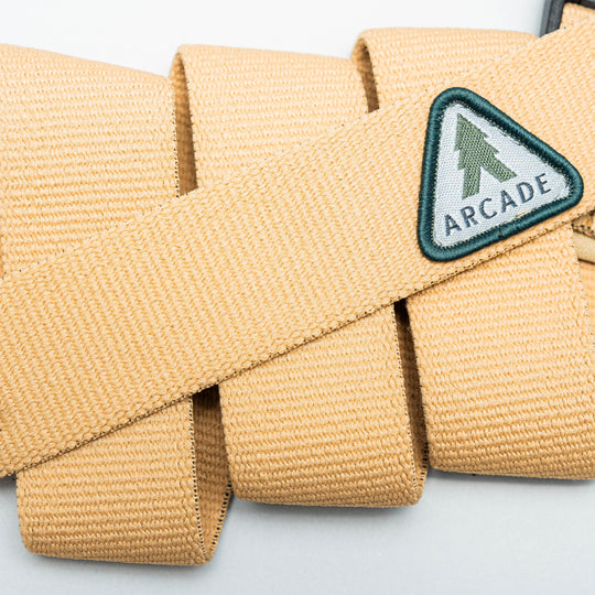 Close-up of the Treeple stretch belt by Arcade Belts, featuring a tan color, plastic buckle, and triangular patch with a green tree and ARCADE text. The low-profile belt is looped on a light background.