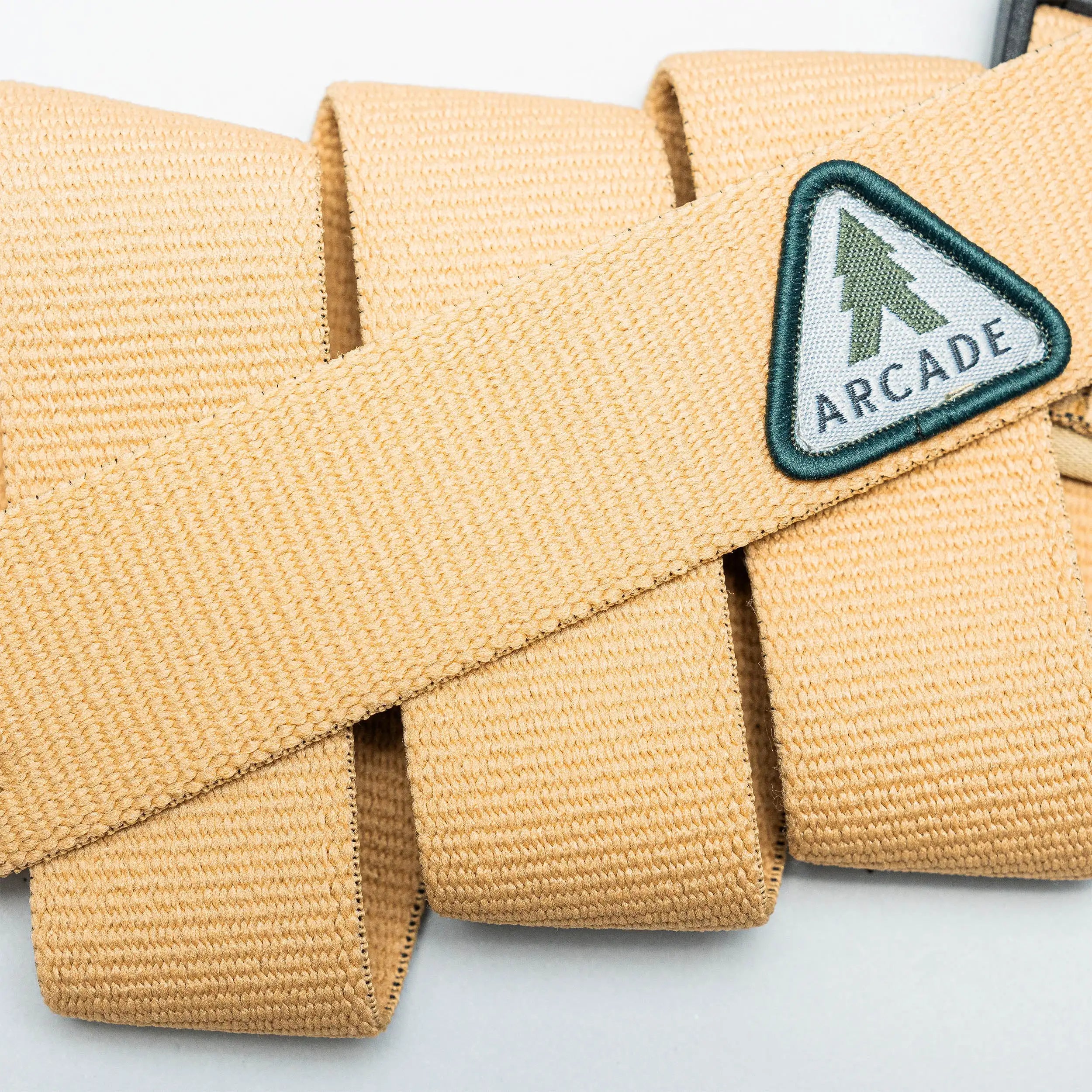 Close-up of the Treeple stretch belt by Arcade Belts, featuring a tan color, plastic buckle, and triangular patch with a green tree and ARCADE text. The low-profile belt is looped on a light background.