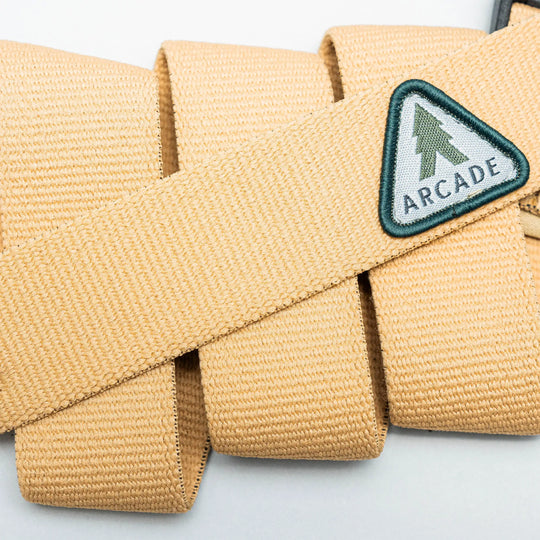 Close-up of the Treeple stretch belt by Arcade Belts, featuring a tan color, plastic buckle, and triangular patch with a green tree and ARCADE text. The low-profile belt is looped on a light background.