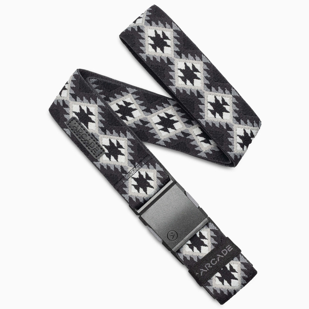 A black and grey Arcade Belts Vernan Kee fabric belt featuring geometric patterns inspired by traditional rugs, with a black plastic buckle and the Arcade brand name displayed near the buckle.