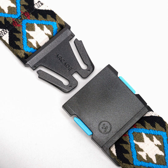 A close-up of the Arcade Belts Vernan Kee low-profile belt with a black plastic buckle marked ARCADE and a small smiling face logo. The strap features a geometric pattern in blue, white, black, and olive green.