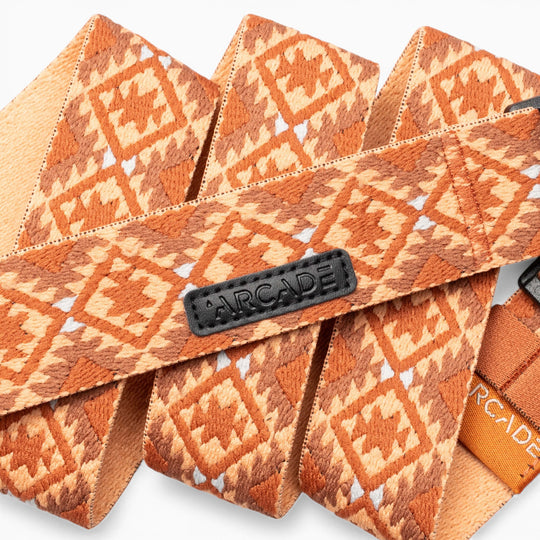 Several orange woven straps with diamond patterns, inspired by Navajo rug designs, are layered together. One strap features a black “Arcade Belts” label, while another displays an orange “Vernan Kee” tag.