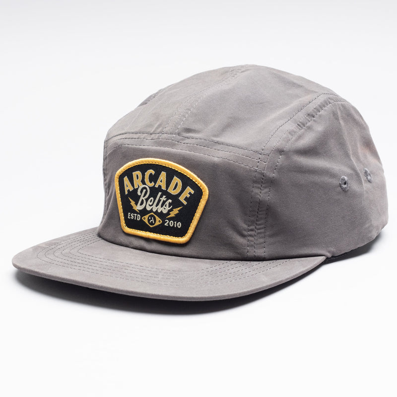 The Arcade Camp Hat is a gray five-panel cap with a flat brim, featuring a black patch on the front with yellow and white Arcade Belts ESTD 2010 text—ideal for pairing with your favorite elastic stretch belt for everyday style.