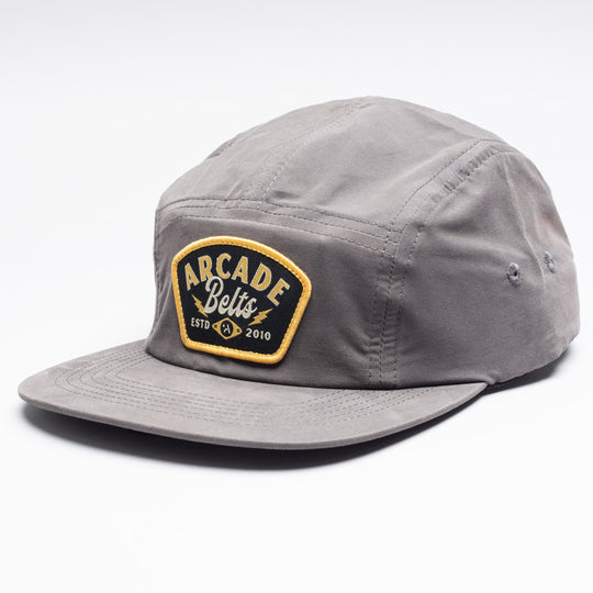 The Arcade Camp Hat is a gray five-panel cap with a flat brim, featuring a black patch on the front with yellow and white Arcade Belts ESTD 2010 text—ideal for pairing with your favorite elastic stretch belt for everyday style.