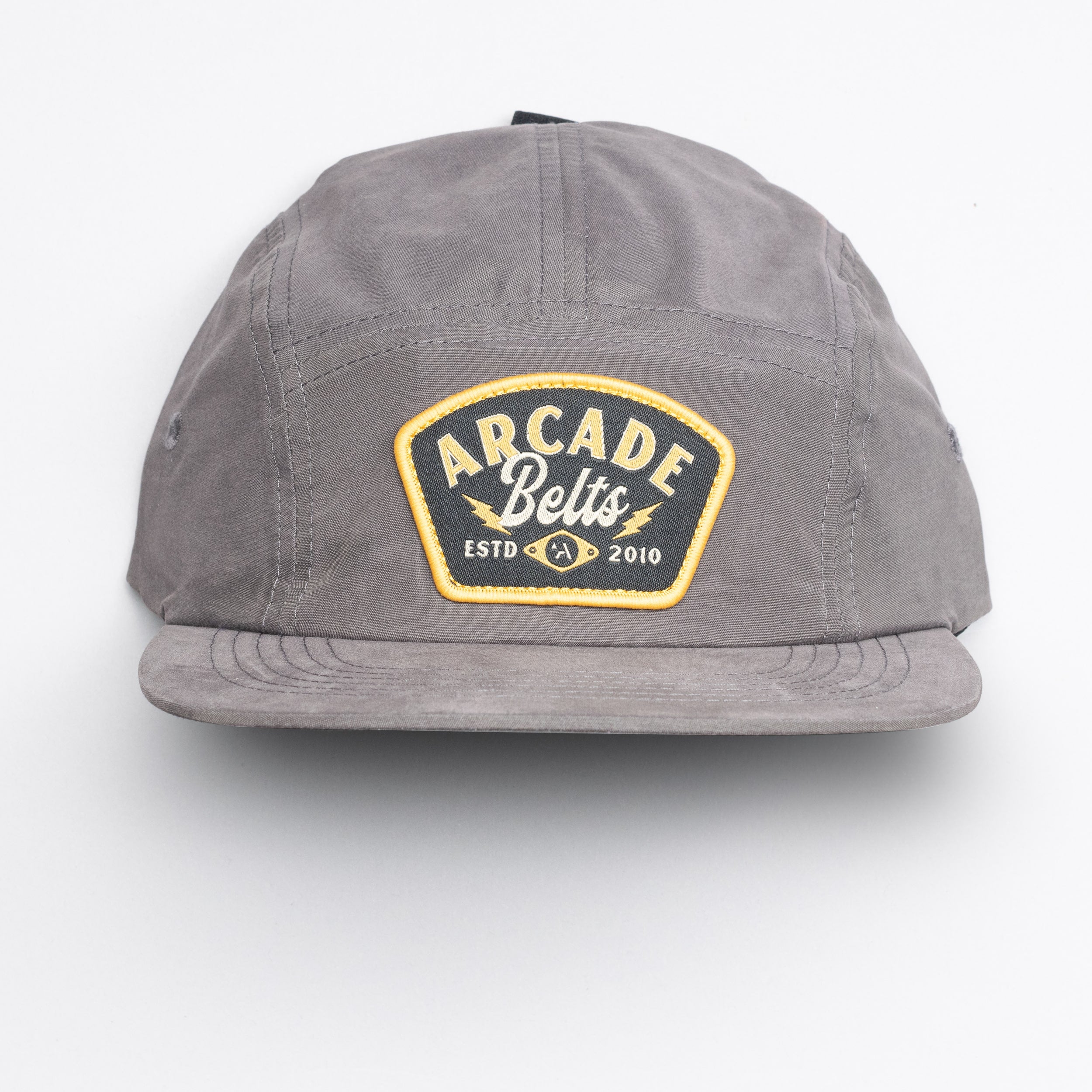The Arcade Camp Hat by Arcade Belts is a gray cap featuring a yellow-bordered front patch with ARCADE Belts ESTD 2010 in white and yellow text, shown on a plain white background—ideal for fans of elastic stretch belt comfort.