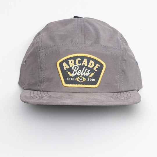 The Arcade Camp Hat by Arcade Belts is a gray cap featuring a yellow-bordered front patch with ARCADE Belts ESTD 2010 in white and yellow text, shown on a plain white background—ideal for fans of elastic stretch belt comfort.
