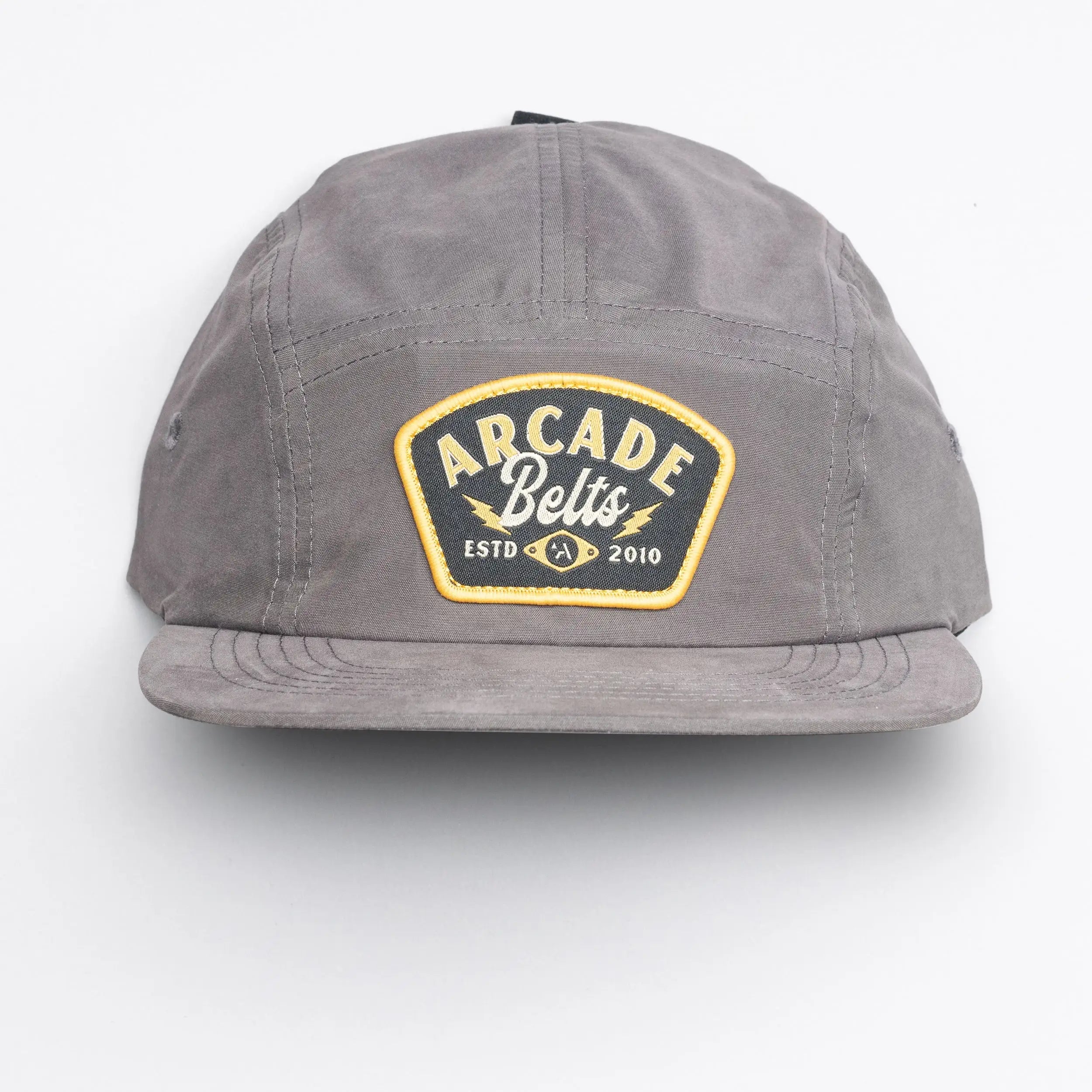 The Arcade Camp Hat by Arcade Belts is a versatile gray baseball cap with a yellow and black "Arcade Belts ESTD 2010" patch, shown against a plain white background.