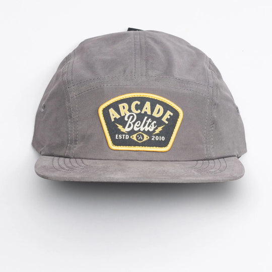 The Arcade Camp Hat by Arcade Belts is a versatile gray baseball cap with a yellow and black "Arcade Belts ESTD 2010" patch, shown against a plain white background.